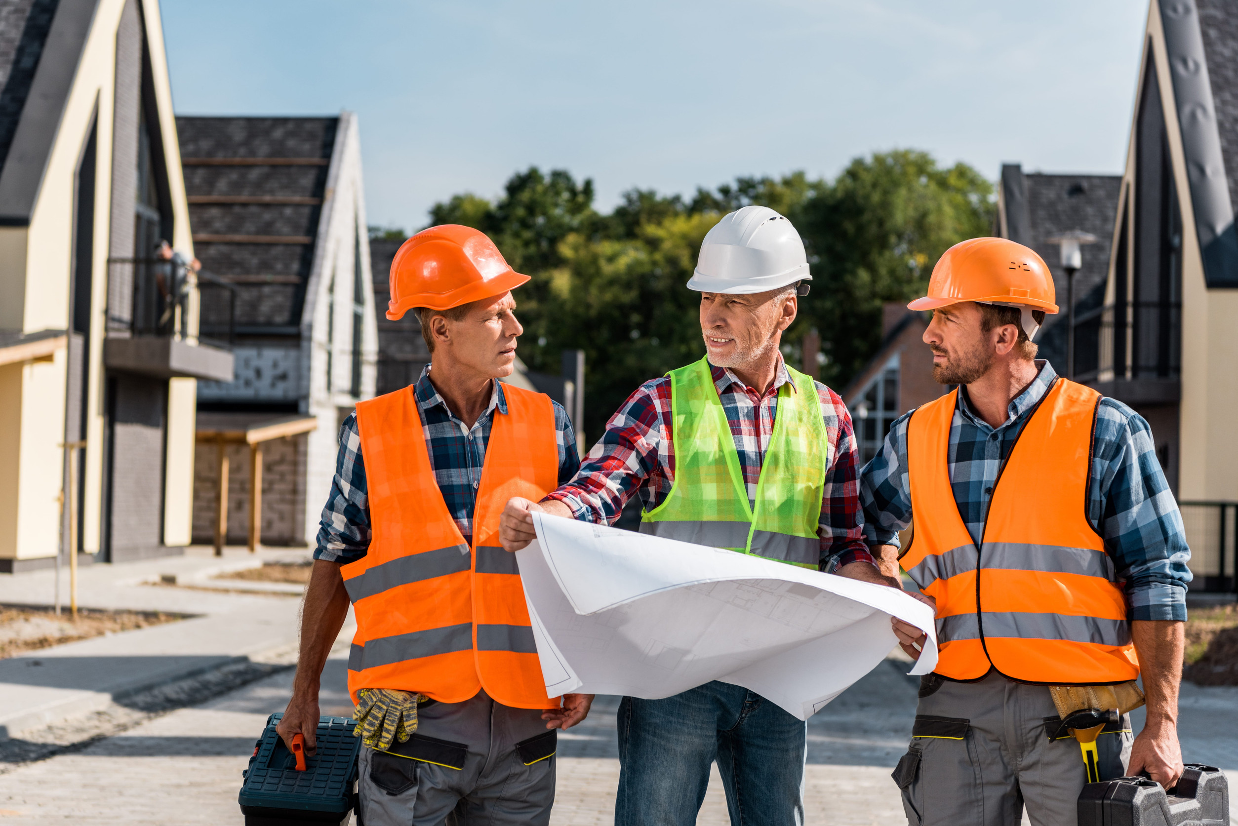 mature builder holding blueprint near coworkers and houses image